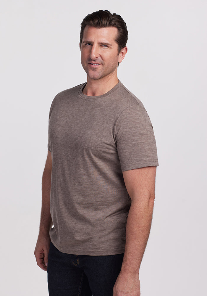 A man wearing a Woolx Endurance Tee in Simply Taupe and dark jeans stands against a plain gray background. He is looking at the camera with a slight smile and a relaxed posture.