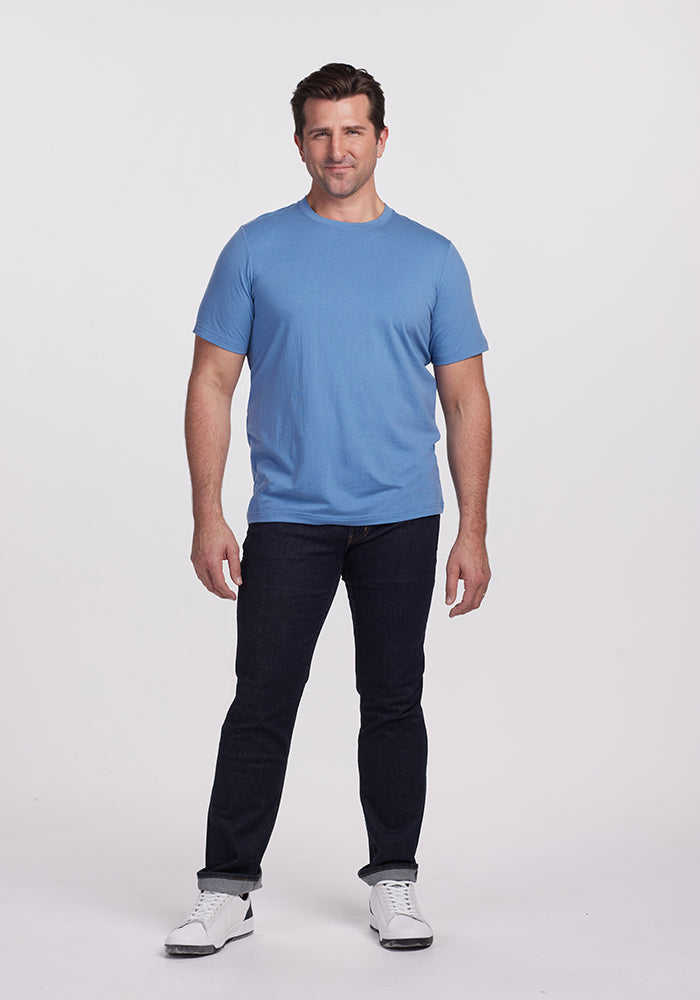 A person wearing a Woolx Endurance Tee in Coronet Blue, along with dark jeans and white sneakers, stands against a plain white background. They have a relaxed posture and a friendly expression.