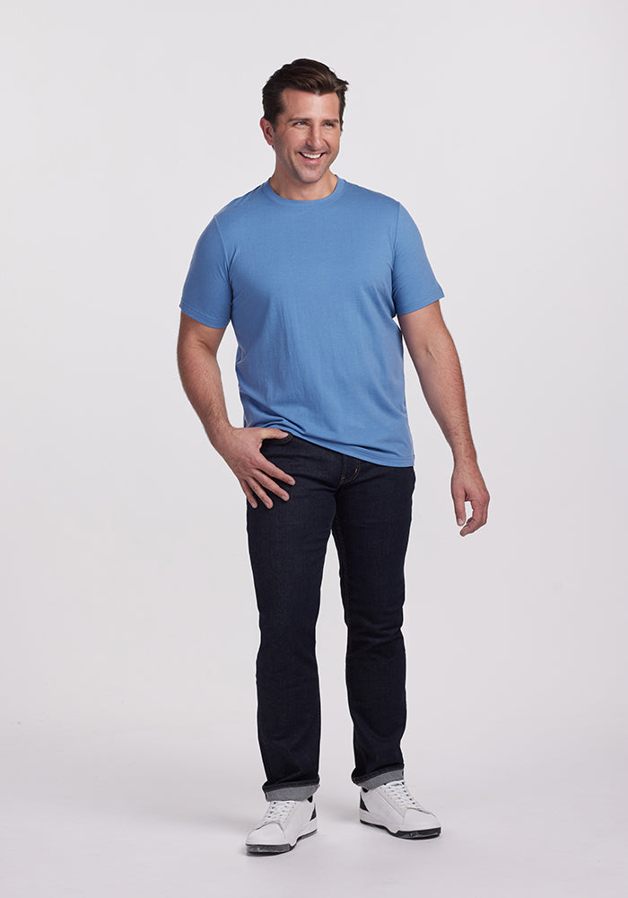 A man wearing the Woolx Endurance Tee in Coronet Blue and dark jeans stands against a plain white background. He sports white sneakers and smiles with his hands relaxed by his sides, possibly enjoying the comfort of Merino Wool performance gear.