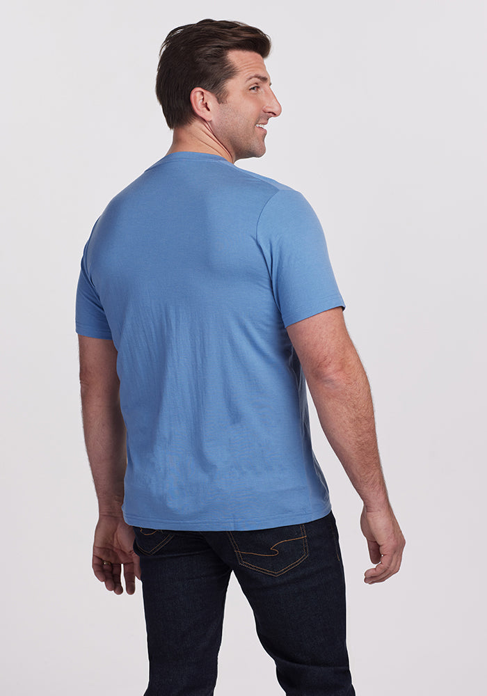 A man dressed in a Coronet Blue Woolx Endurance Tee and dark jeans stands facing away, turning slightly to his right against a plain white background.