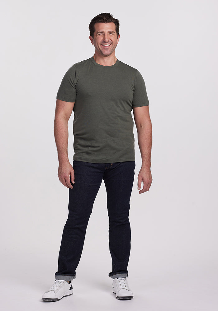 A person is standing and smiling, wearing a vibrant Endurance Tee in Deep Fern by Woolx, dark jeans, and white sneakers against a plain white background.