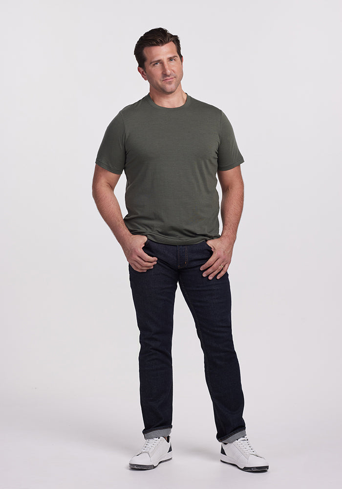 A person stands against a plain background wearing an Endurance Tee in Deep Fern, dark jeans, and white sneakers. The person has short brown hair and their hands are casually tucked into their jeans pockets. The tee is by Woolx.