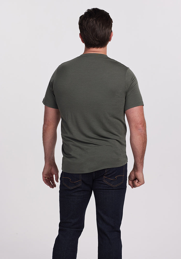 A man with short dark hair wears the Woolx Endurance Tee in Deep Fern and dark blue jeans. The outfit, featuring performance gear, ensures comfort as he stands with his back to the camera against a plain white background.