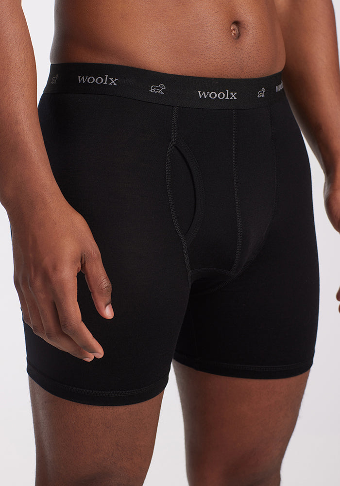 A person wearing Woolx Reaction Boxer Briefs in black stands against a plain white background. The waistband displays the brand name in white font. This comfortable underwear highlights exceptional design and fit, positioning itself as a leading choice for performance boxers.