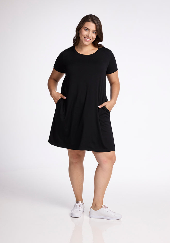 A woman is standing and smiling in the Georgie Dress - Black by Woolx, with her hands in the side pockets. She is wearing white sneakers and is posed against a plain white background. 