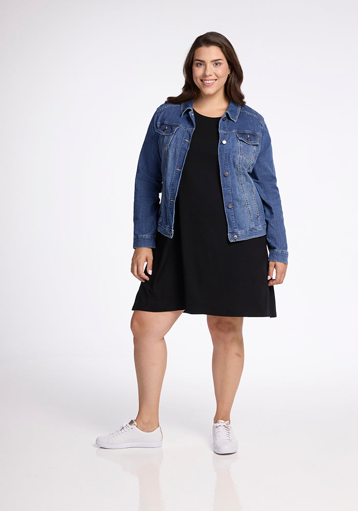 A woman is standing and smiling at the camera, wearing a blue denim jacket over a relaxed fit Georgie Dress - Black by Woolx, paired with white sneakers. The background is plain white.