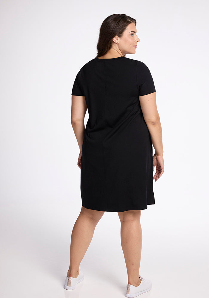 A person with long brown hair is shown from the back, standing and wearing a simple black Woolx Georgie Dress with short sleeves. They are also wearing white sneakers, standing on a white background. The relaxed fit of the dress adds to the casual elegance of the ensemble.