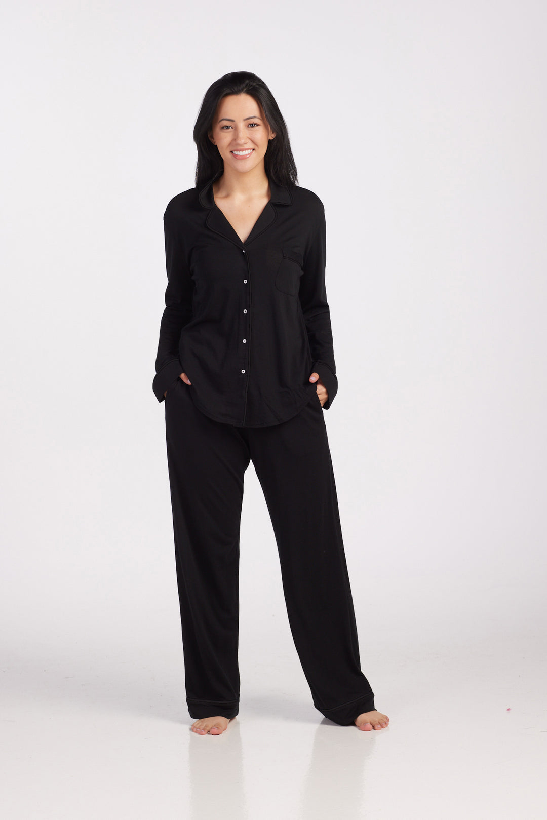 A person wearing the Harper FeatherTouch® Pajama Set in Black from Woolx stands barefoot against a plain white background, smiling with hands in their pockets. Renowned for its comfortable sleepwear, this set is ideal even during night sweats.