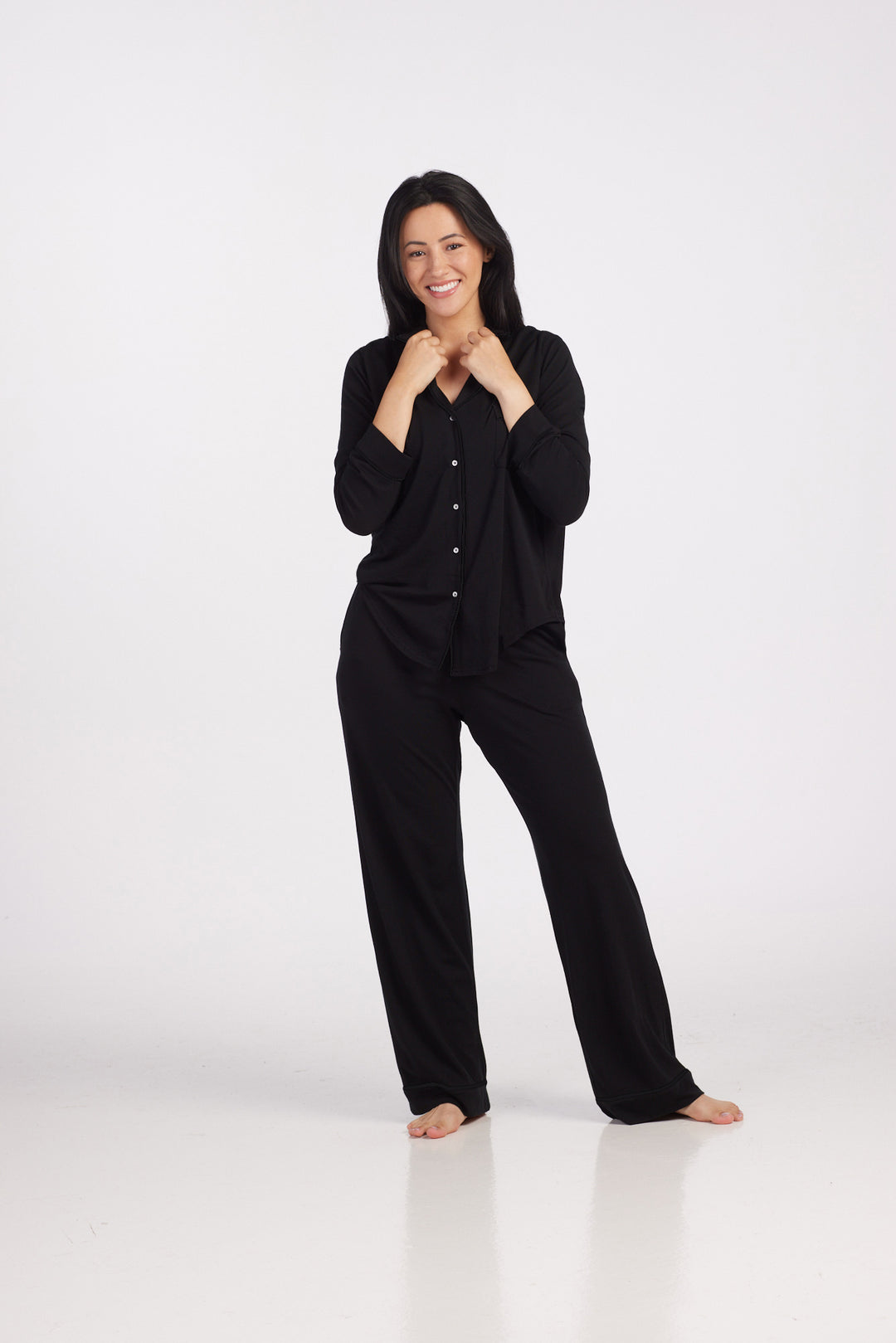 A person with long dark hair is smiling and standing barefoot on a white background. Clad in the Harper FeatherTouch® Pajama Set - Black from Woolx, they sport a black button-up shirt and pants designed for comfort. Their hands are clasped at the collar, exuding relaxed elegance ideal for combating night sweats.