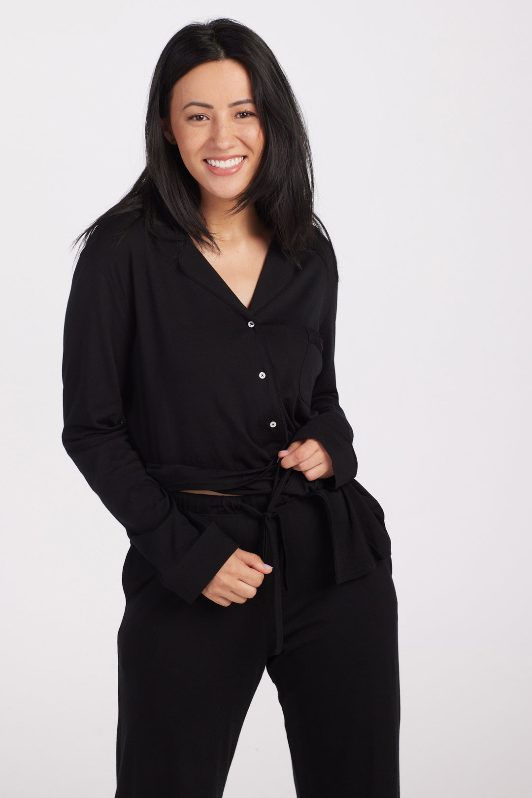 A person with long black hair smiles at the camera, dressed in the Harper FeatherTouch® Pajama Set - Black by Woolx, against a plain white background. 