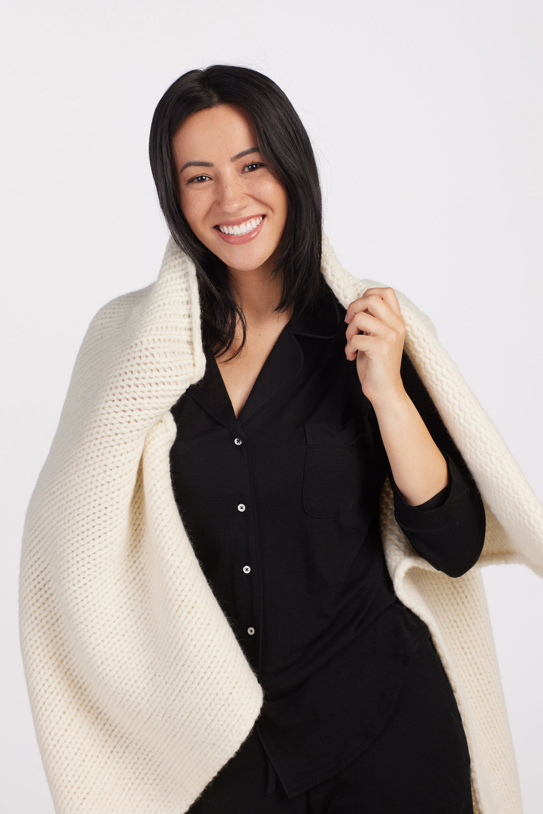 A person with long dark hair and a bright smile is wrapped in a cozy cream-colored knit blanket. Wearing the Harper FeatherTouch® Pajama Set in black from Woolx against a plain white background, they radiate comfort and ease.