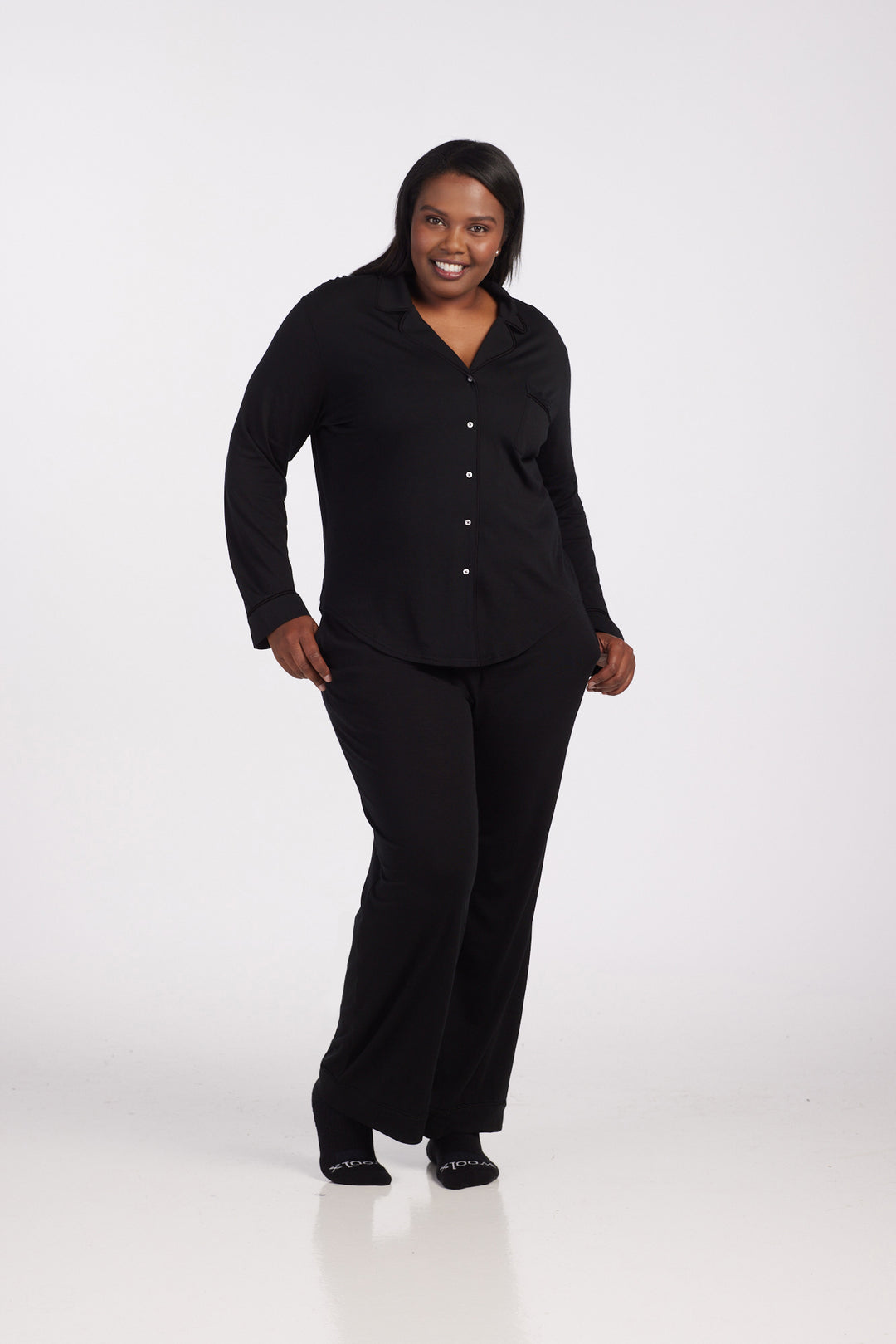 A person dressed in a black Harper FeatherTouch® Pajama Set by Woolx, consisting of a button-up shirt and pants, stands against a plain white background, smiling and walking forward. 