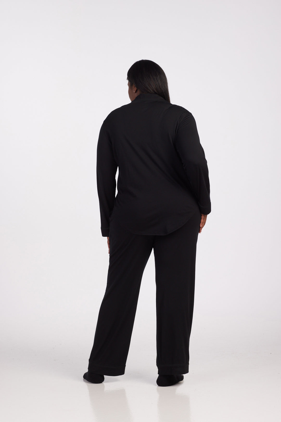 A person stands with their back to the camera, dressed in the Harper FeatherTouch® Pajama Set - Black from Woolx. The background is neutral and minimal, creating a serene vibe.