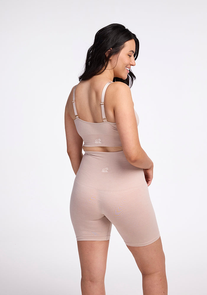 A smiling woman with long, dark hair is posing sideways, looking over her shoulder. She is wearing beige Marley Shapewear Shorts by Woolx and a seamless bra crafted from lightweight merino wool. The background is plain white.