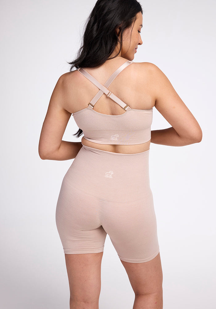 A woman is depicted from behind in beige, form-fitting activewear. The outfit features a crop top with crossed back straps paired with Woolx Marley Shapewear Shorts that are high-waisted. She has long dark hair and is standing against a plain, light-colored background.