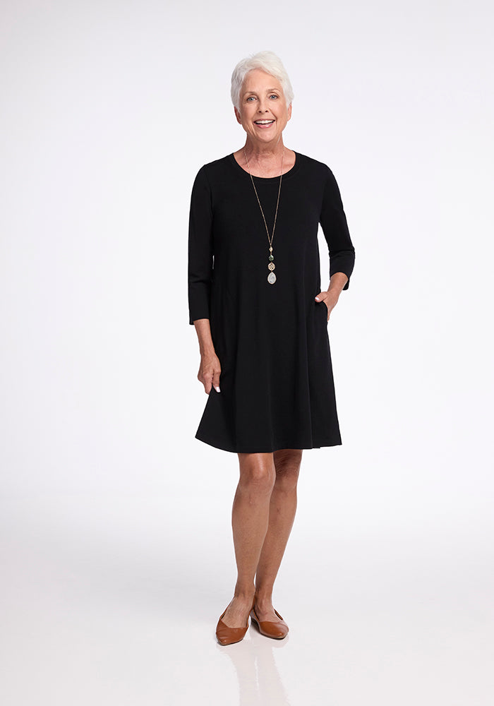 An older woman with short white hair is wearing a sleek Rissa A Line Dress - Black from Woolx, expertly crafted from merino wool for comfortable travel. Her flat brown shoes perfectly complement the outfit as she stands against a plain white background, smiling with one hand resting in her pocket and the other by her side. A long necklace adds to her layer-friendly ensemble.