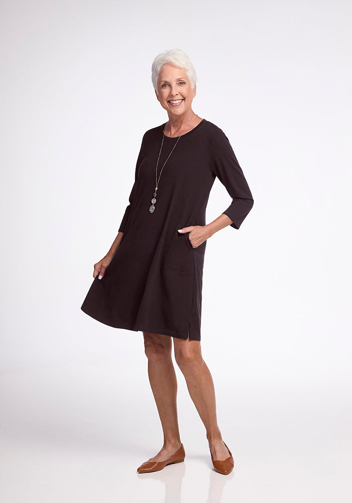 An older woman with short white hair is beaming as she stands in a Woolx Rissa A Line Dress in French Roast, featuring three-quarter sleeves and convenient pockets. The dress's A-line silhouette beautifully enhances her figure, pairing perfectly with her long necklace and brown flats. She is set against a plain white background.