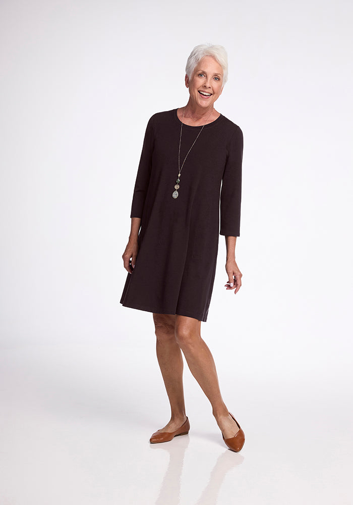 An elderly woman with short white hair is smiling as she stands in a light, empty room. She's elegantly dressed in a long-sleeve Rissa A Line Dress - French Roast by Woolx, complemented by her brown flats.