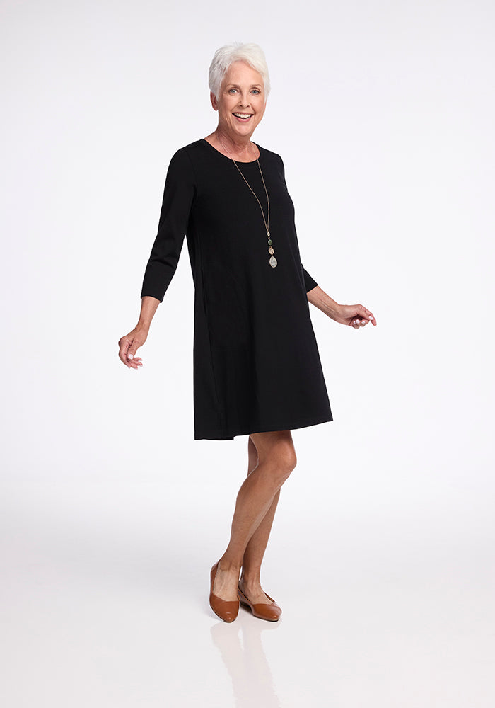 A woman with short white hair joyfully poses in the Rissa A Line Dress - Black by Woolx, and brown shoes against a plain white background. The dress, perfect for travel thanks to its layering flexibility, is accessorized with a long necklace, adding to her charming style.