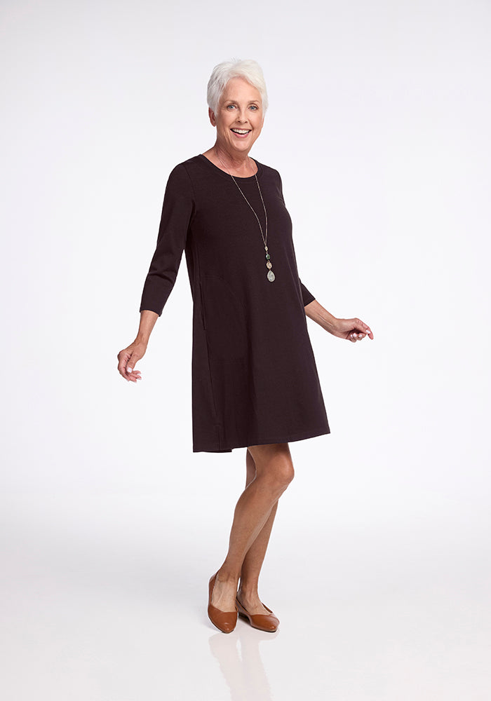 A confident older woman with short gray hair smiles warmly, showcasing her Rissa A Line Dress in French Roast by Woolx. She steps forward with her brown shoes and wears a long necklace, striking a pose against the plain white backdrop.