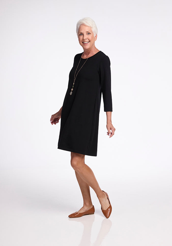 An older woman with short white hair smiles while wearing the black Rissa A Line Dress from Woolx and a pair of brown flats. She stands with her weight on one leg, creating a dynamic pose against a plain white studio backdrop.