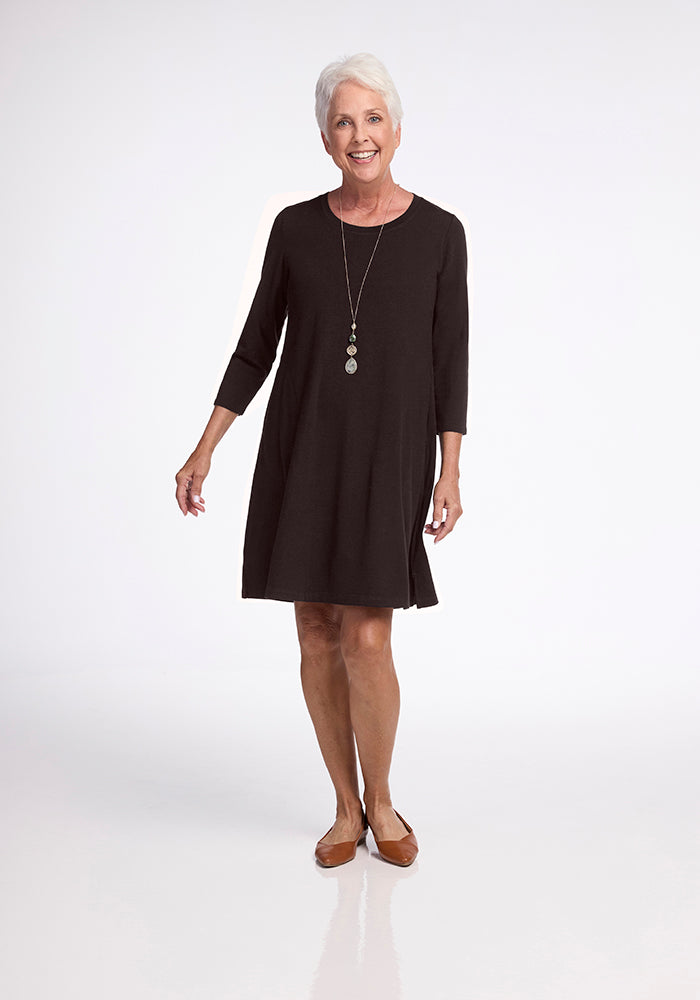 An older woman with short gray hair smiles while wearing the knee-length Rissa A Line Dress in French Roast from Woolx and brown shoes. She stands against a plain white background, accessorized with a long necklace featuring two circular pendants.