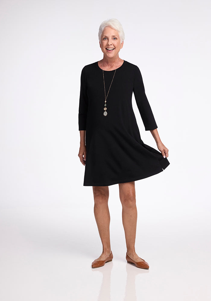 A smiling older woman with gray hair wears a layer-friendly Rissa A Line Dress in black by Woolx, paired with brown flat shoes. She stands on a plain white background, holding the hem of her travel dress with one hand. Her outfit is completed by a long necklace featuring circular pendants.