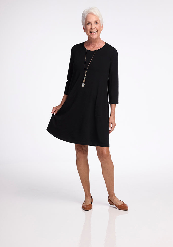 An elderly woman with short white hair is smiling as she poses against a plain white background, holding the hem of her knee-length Rissa A Line Dress in black from Woolx. She complements her stylish travel outfit with brown flats and a long necklace, offering both comfort and versatility that's perfect for layering on any trip.