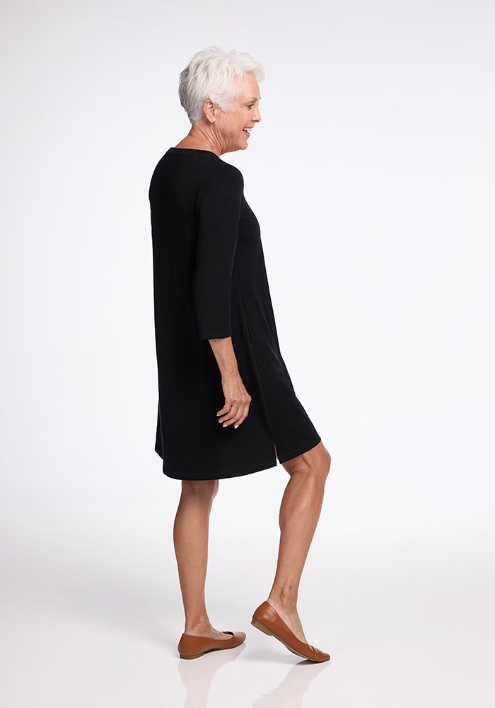 An older adult with short white hair stands sideways, wearing the Rissa A Line Dress - Black from Woolx and a pair of brown flats. They are smiling as they walk against a plain white background.