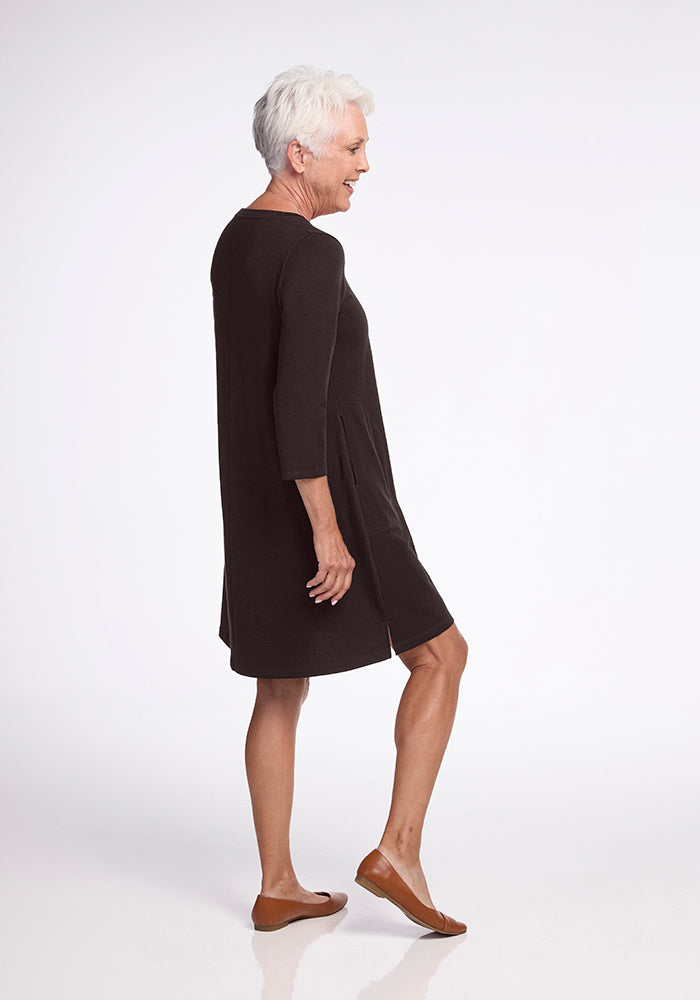 An older woman with short white hair is wearing the Woolx Rissa A Line Dress in French Roast and brown flats, standing sideways and smiling against a plain white background.