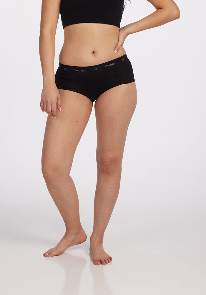 A person wearing a black top and Woolx's Lila Boy Shorts - Black, which are Woolmark Certified athletic underwear, stands against a plain white background. Their left hand rests on their thigh in a pose that embodies comfort, and they are barefoot.