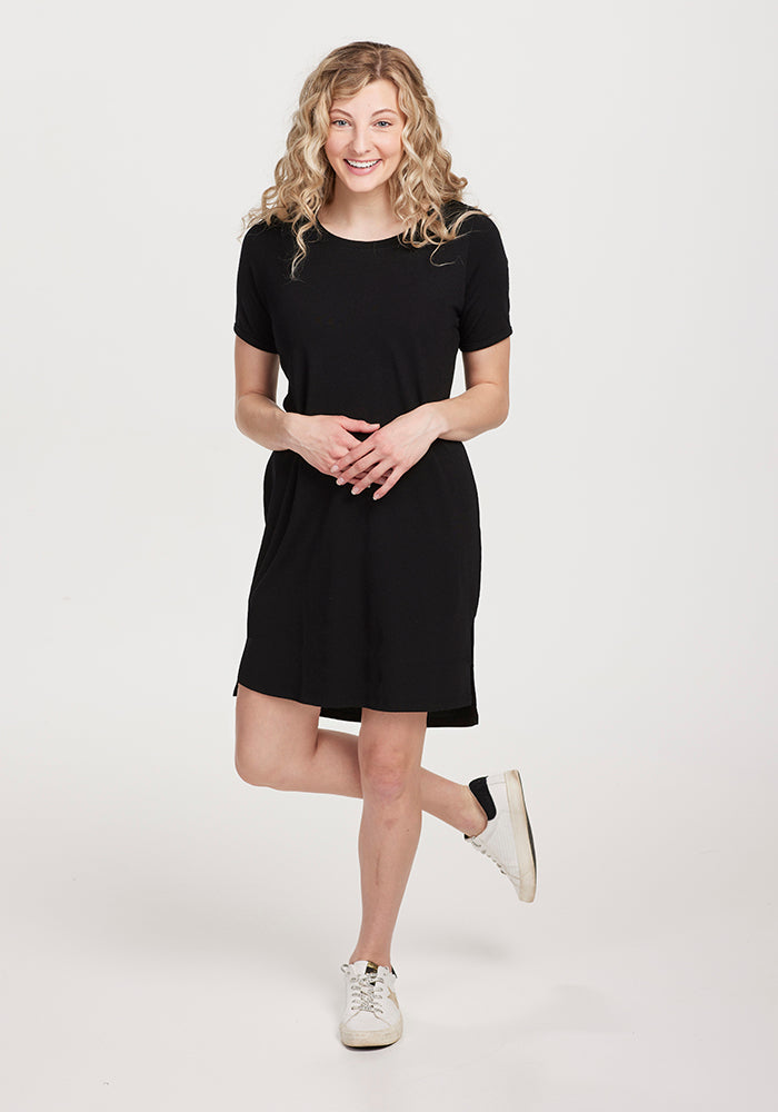 A person with curly hair is smiling while standing in the Lexie Dress & Cover-up OLD by Woolx, a black knee-length dress, paired with white sneakers against a plain background.