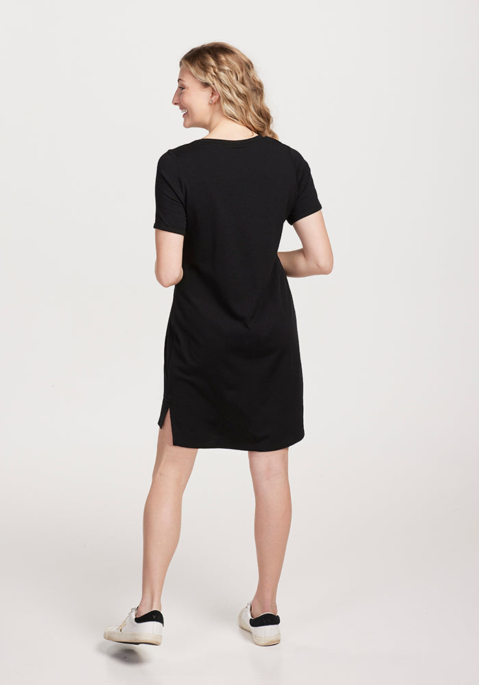 A woman with long, wavy blonde hair stands facing away in a classic silhouette, wearing the Lexie Dress & Cover-up OLD from Woolx, crafted in black Merino wool with short sleeves and paired with white sneakers. She is set against a light gray background.