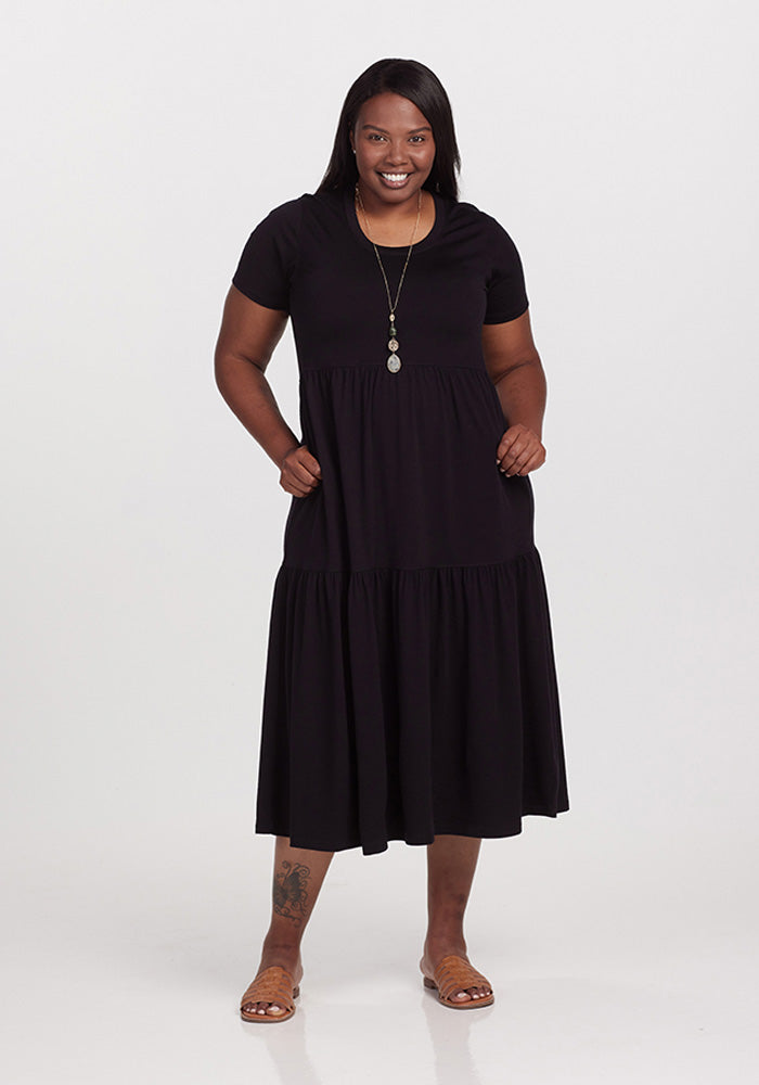 A person is smiling and wearing a Woolx Lucia Dress in Black, crafted from merino wool, paired with sandals. They have their hands tucked into the dress pockets, with a necklace subtly visible. The plain white background accentuates the chic summer vibe of this dress perfectly. 