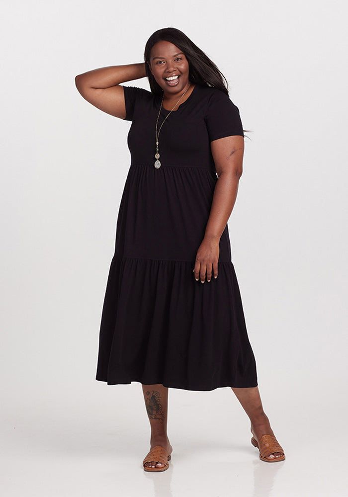 A person dressed in a Woolx Lucia Dress - Black and brown sandals stands smiling against a plain background. They are posing with one hand on their head, complemented by a long necklace, creating an ideal summer dress look.