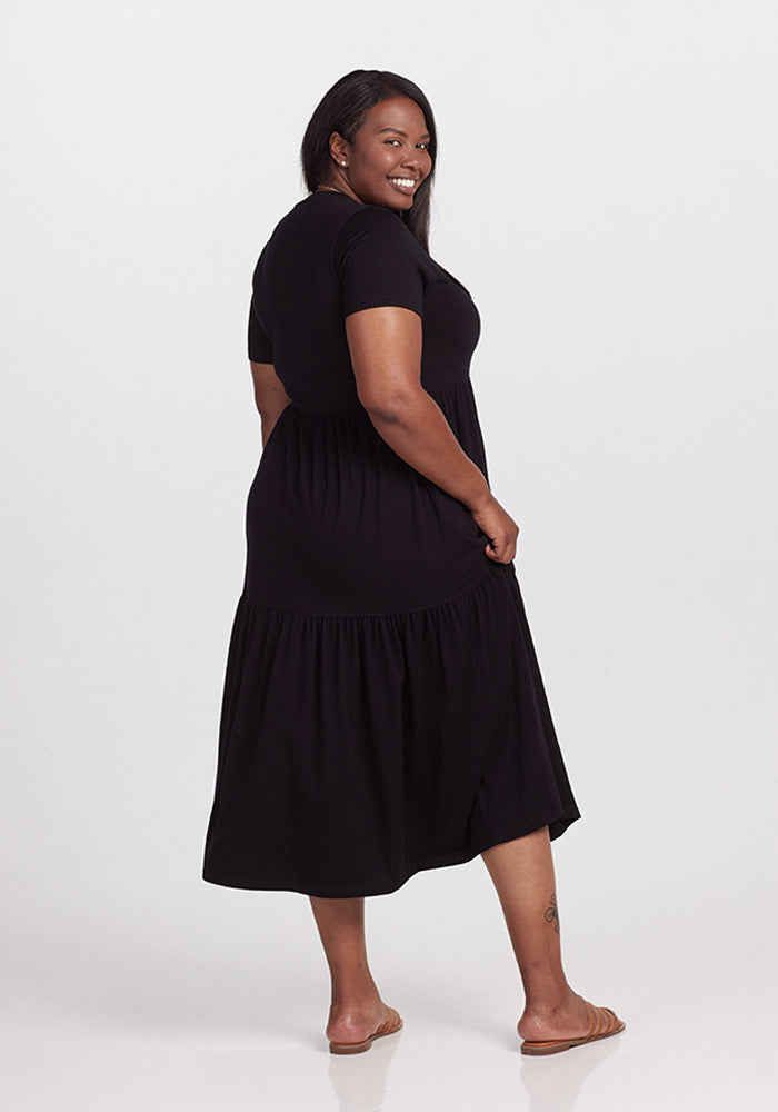 A person wearing a black Woolx Lucia dress and sandals stands against a plain white background. They are smiling and looking over their shoulder, capturing an effortless style perfect for warm days.