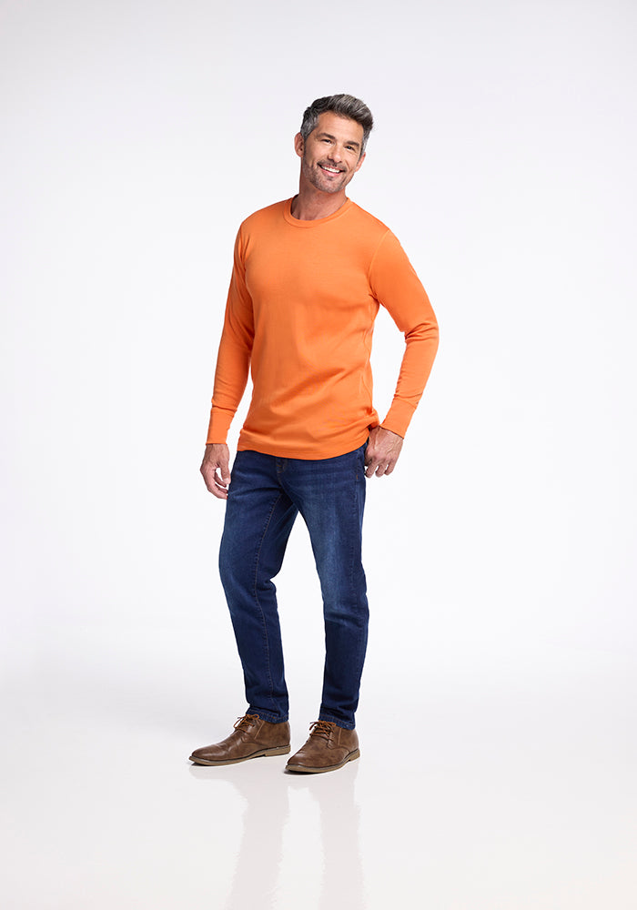 A man stands against a plain white background, smiling and looking slightly to the side. He is wearing an orange Woolx Explorer Base Layer shirt, dark blue jeans, and brown shoes. His right hand rests on his hip, and his left hand hangs loosely by his side, ready for outdoor adventures.