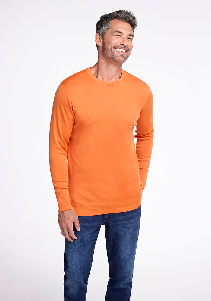 A smiling man with gray hair and a short beard is standing against a white background. He is wearing the Woolx Explorer Base Layer under his orange long-sleeve shirt and blue jeans, with his hands in his pockets, ready for outdoor adventures.