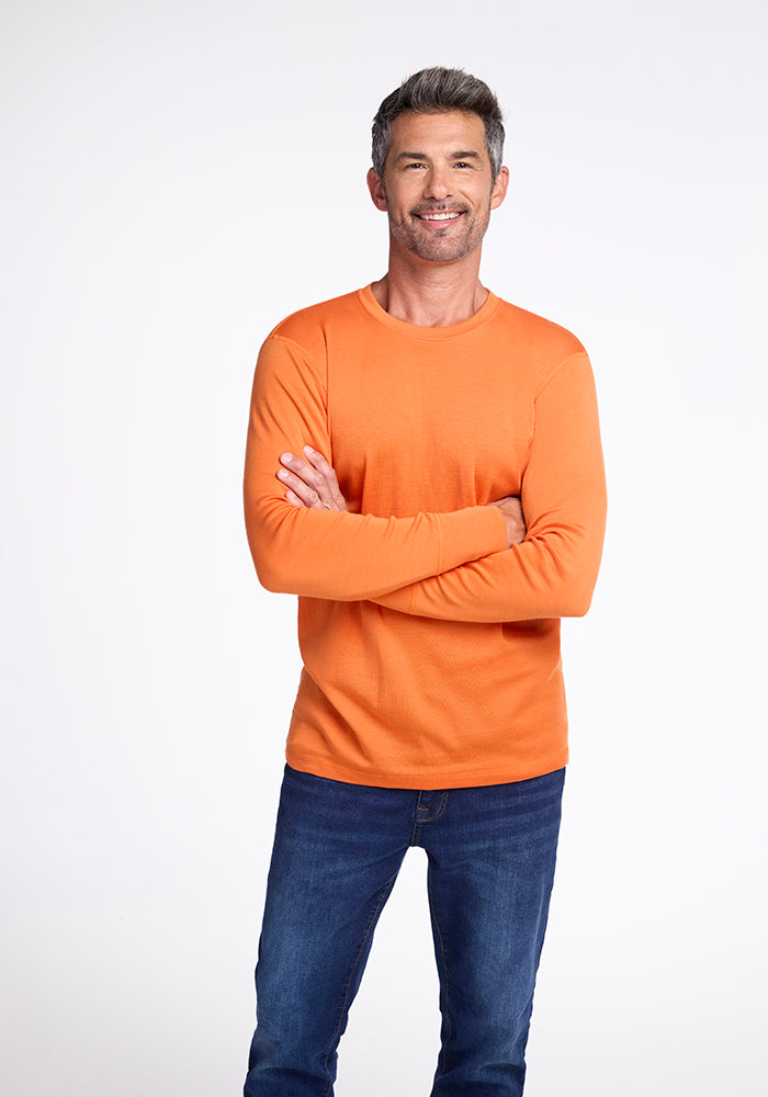 A man with short, gray hair is smiling and posing with his arms crossed. He is wearing an orange Woolx Explorer Base Layer and blue jeans. The background is plain white. 