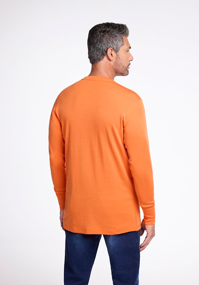 A man with short gray hair is standing and facing away from the camera, showcasing his long-sleeved, bright orange Explorer Base Layer from Woolx paired with dark blue jeans. The plain white background draws full attention to the man and his vibrant attire.