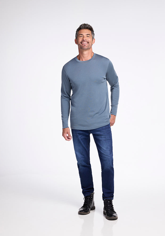 A man stands against a plain white background, wearing the Woolx Explorer Base Layer, a long-sleeve, blue-gray midweight performance shirt. He pairs it with dark blue jeans and black boots. He is smiling and appears to be walking forward with one foot slightly lifted, ready for his next outdoor adventure.