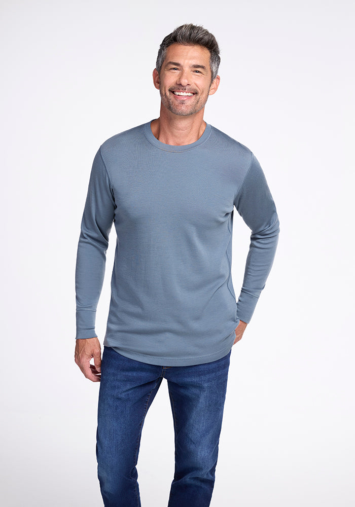 A man with short, graying hair is smiling and standing against a plain white background. He is wearing a light blue Explorer Base Layer by Woolx and dark blue jeans. His right hand is relaxed by his side while his left thumb is hooked into his jeans pocket. 