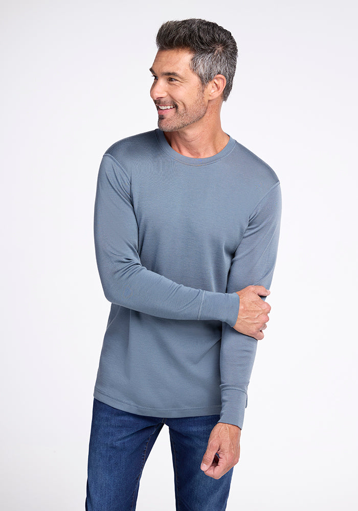 A man with short grey hair, a salt-and-pepper beard, and a friendly expression is wearing a blue Woolx Explorer Base Layer and blue jeans. He is standing against a plain white background, smiling, and looking to his left while holding his right elbow with his left hand.