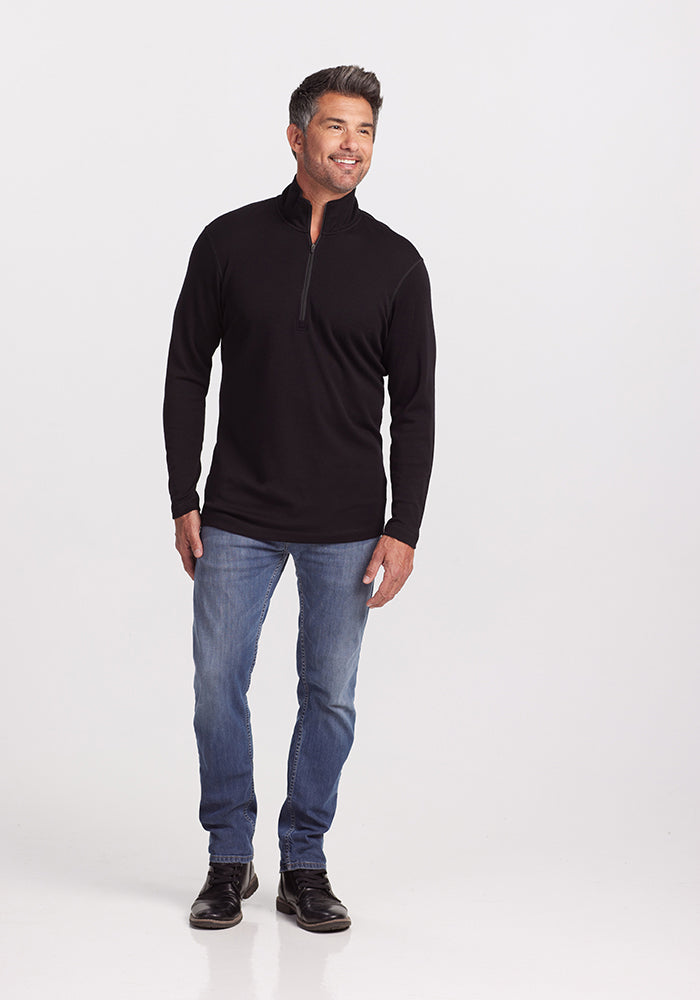 A man with short, dark grey hair is wearing the Woolx Explorer 1/4 Zip in black, made from ultra-soft midweight fabric. He pairs it with blue jeans and black shoes. Standing casually with one hand at his side and the other slightly bent, he smiles and looks off to the side against a plain white background.