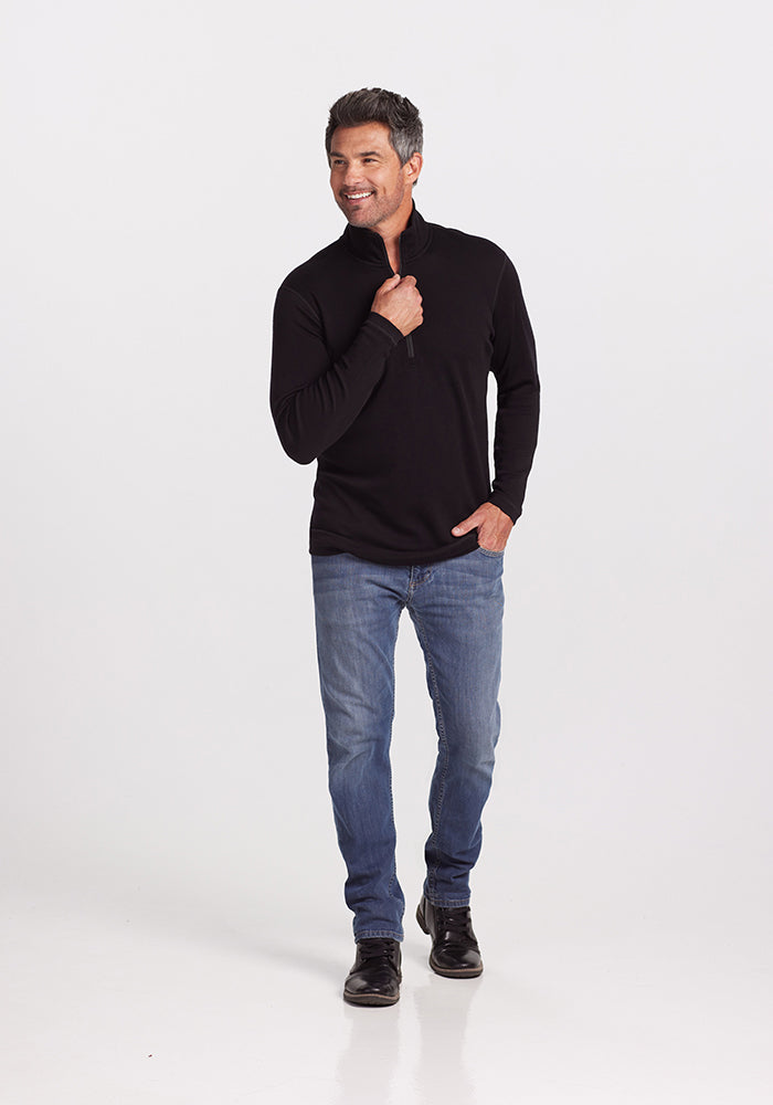 A man is standing against a plain white background. He is wearing the Explorer 1/4 Zip - Black from Woolx, which is crafted from luxurious Merino wool, along with blue jeans and black shoes. He is smiling, one hand holding the zipper near his chest, and the other hand resting in his jeans pocket.