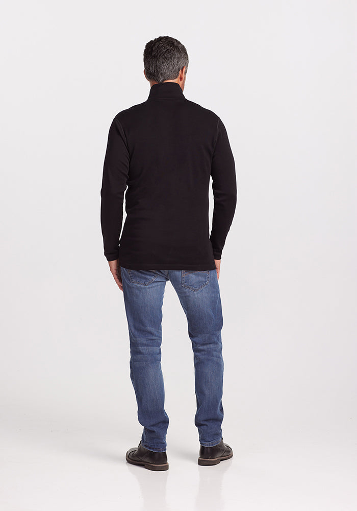 A man with gray hair stands facing away from the camera. He is wearing a Woolx Explorer 1/4 Zip in black, made of midweight fabric, blue jeans, and dark shoes. The background is plain white.