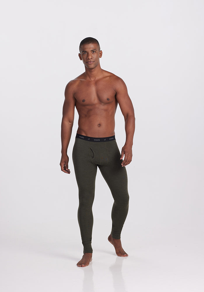 A muscular man poses barefoot against a plain white background, wearing Woolx's Backcountry Leggings in Dark Moss made from performance fabric. He confidently stands shirtless with a neutral expression and his hands by his sides, prepared for cold weather adventures.