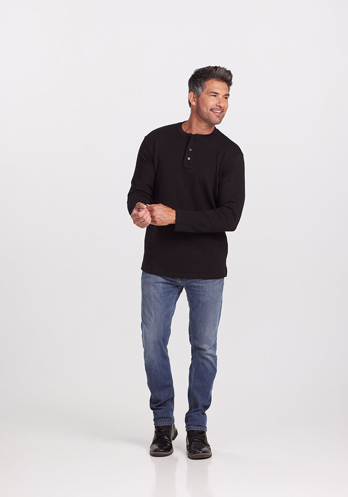 A man stands against a plain white background, wearing the Jackson Henley - Black from Woolx, a black long-sleeve henley shirt made from midweight Australian Merino wool. He pairs it with blue jeans and black shoes. Smiling warmly, he looks slightly to his left with his hands held in front of him.