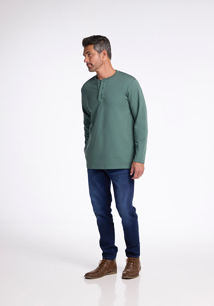 A man in a green Jackson Henley - Duck Green shirt by Woolx, dark blue jeans, and brown leather shoes stands against a plain white background. He is looking to his left with his hands loosely by his sides.