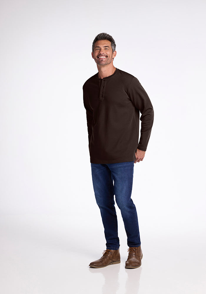 A man with grey hair is smiling and standing against a plain white background. He is wearing a dark brown heavyweight Jackson Henley from Woolx, blue jeans, and brown lace-up boots. His right hand is clenched into a fist, and he appears relaxed and happy.